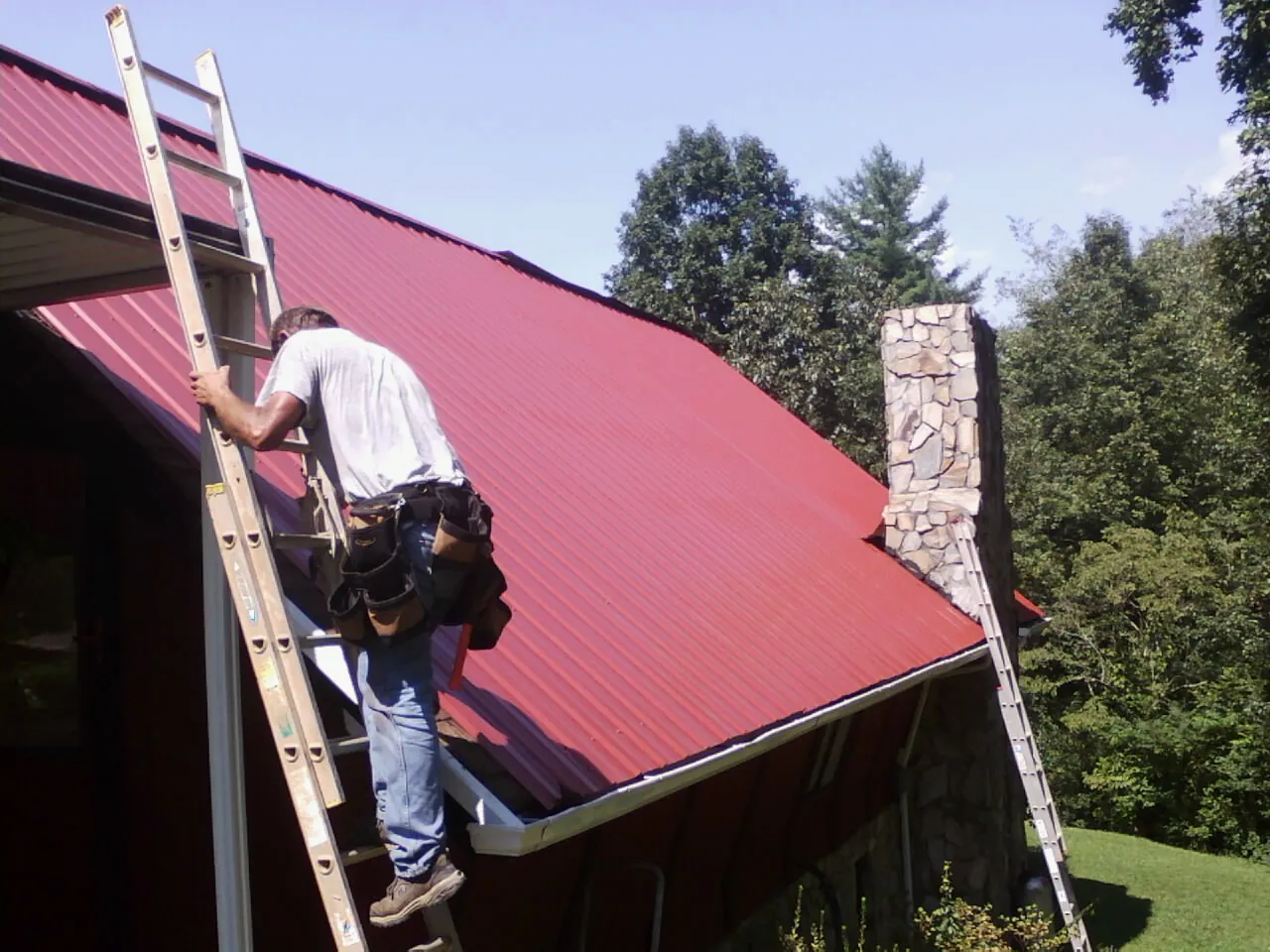 Roofer on a ladder beside a red metal roof and stone chimney — Soffit & Fascia Repair by Roofing Brothers in Golden Valley